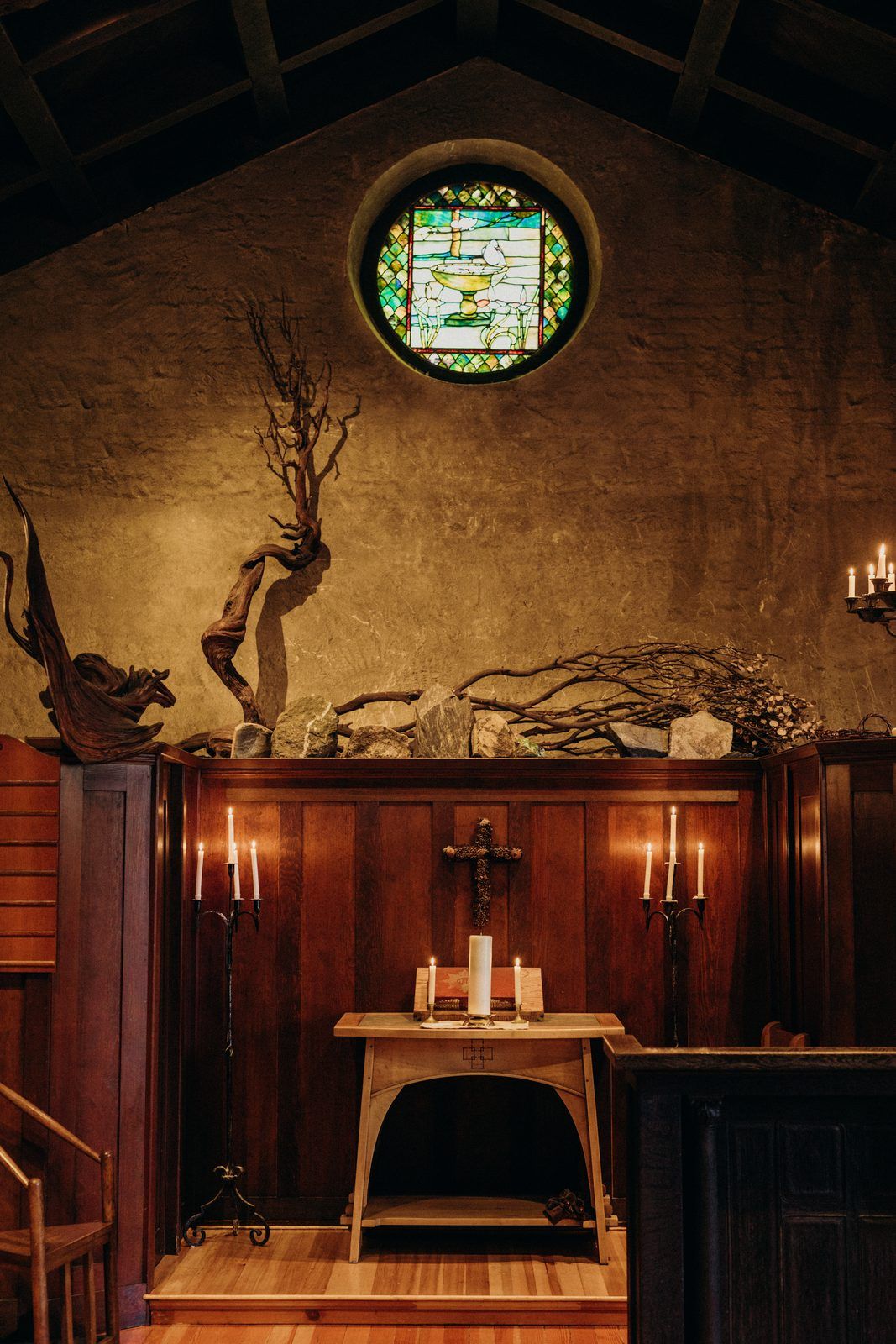 The altar at the end of the church: a small wooden table holding a lit candle and an open book, candelabra on either side, and a round stained glass window in the wall above.