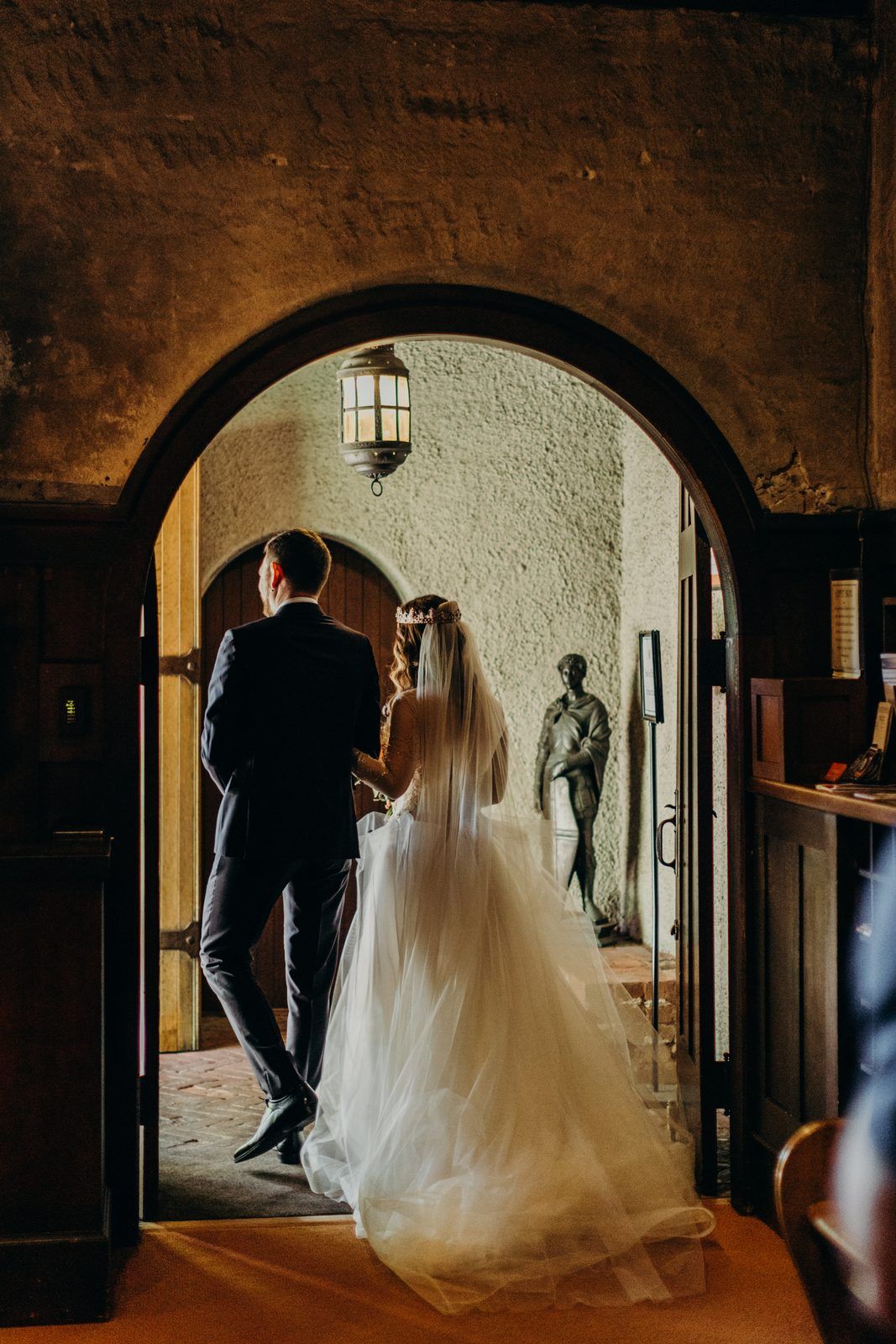 The couple seen from behind walking out through a stone arched doorway into the courtyard after the ceremony.