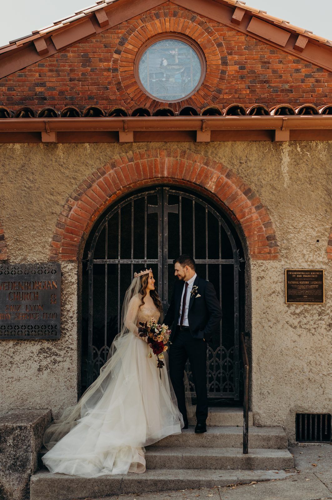 Rena and Colin standing together on the steps of the Swedenborgian Church entrance, a brick arched gateway under a round window.