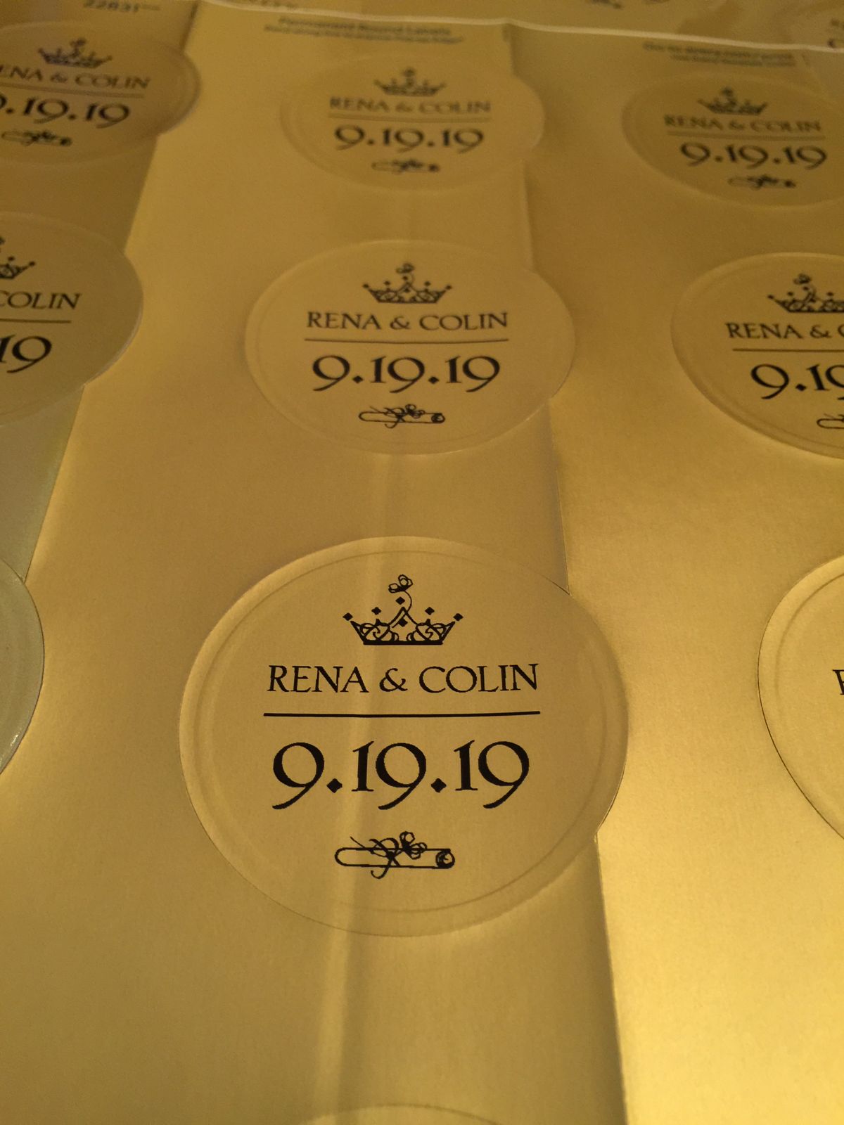 A sheet of round clear labels, each printed with a small crown, the words Rena & Colin, the date 9.19.19 and a little scroll.