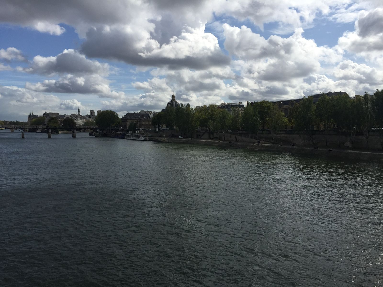 Heavy white and grey clouds over the Seine in Paris, the river dark below and trees and stone buildings along the far bank.