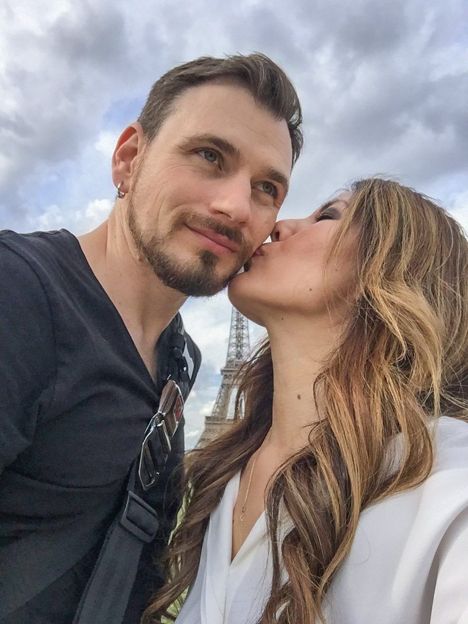 Rena kissing Colin on the cheek in a selfie, the Eiffel Tower behind them under a cloudy sky.