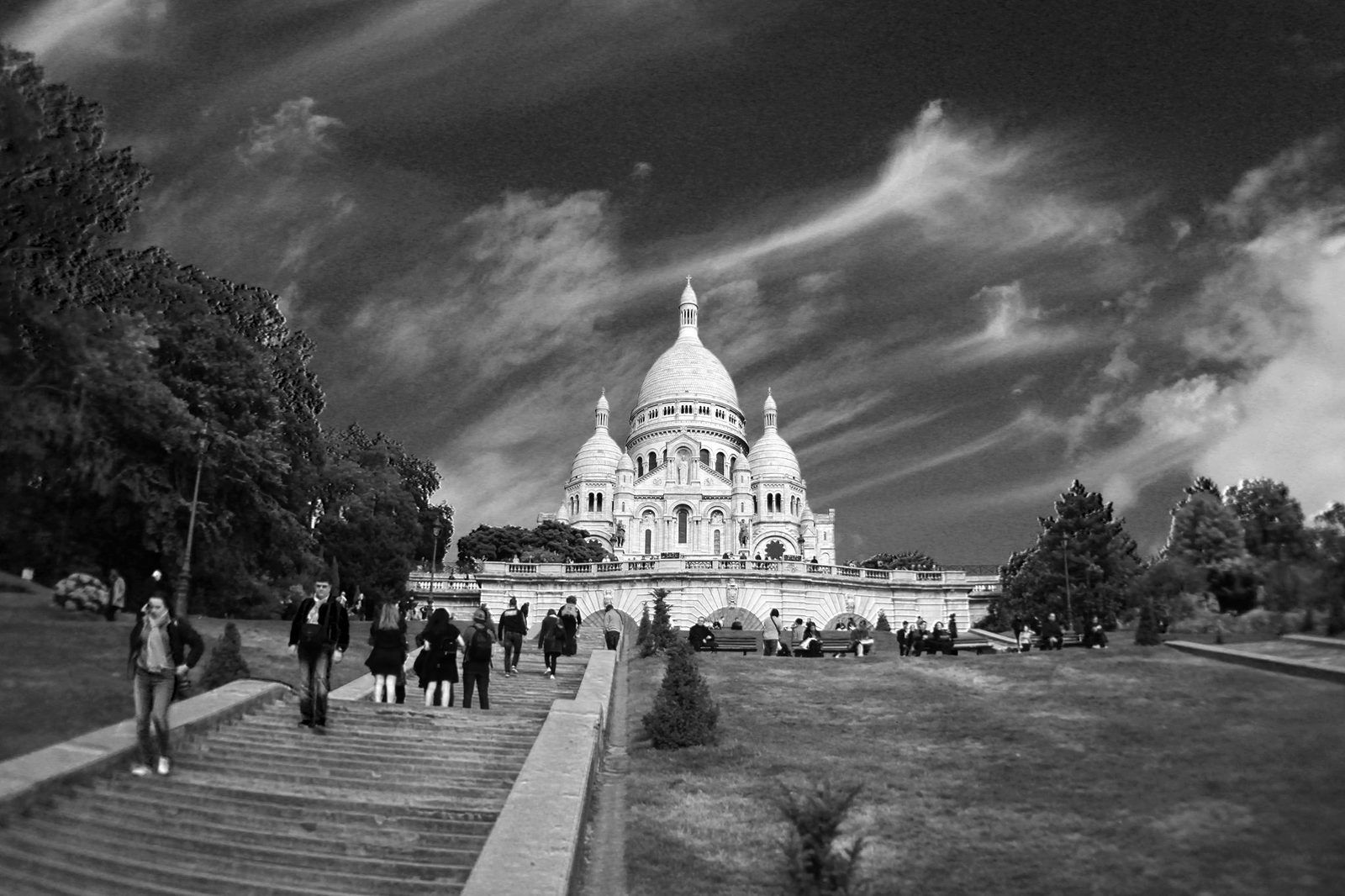 The white domes of the Sacre-Coeur in Montmartre seen up a flight of steps and a lawn, under a dramatic sky, in black and white.