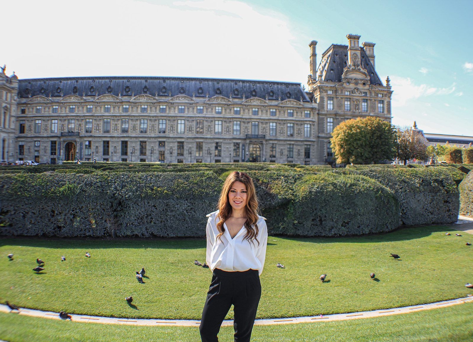 Rena standing in a white blouse and black trousers on a lawn in the Tuileries with pigeons around her, the long stone wing of the Louvre and a clipped hedge behind.