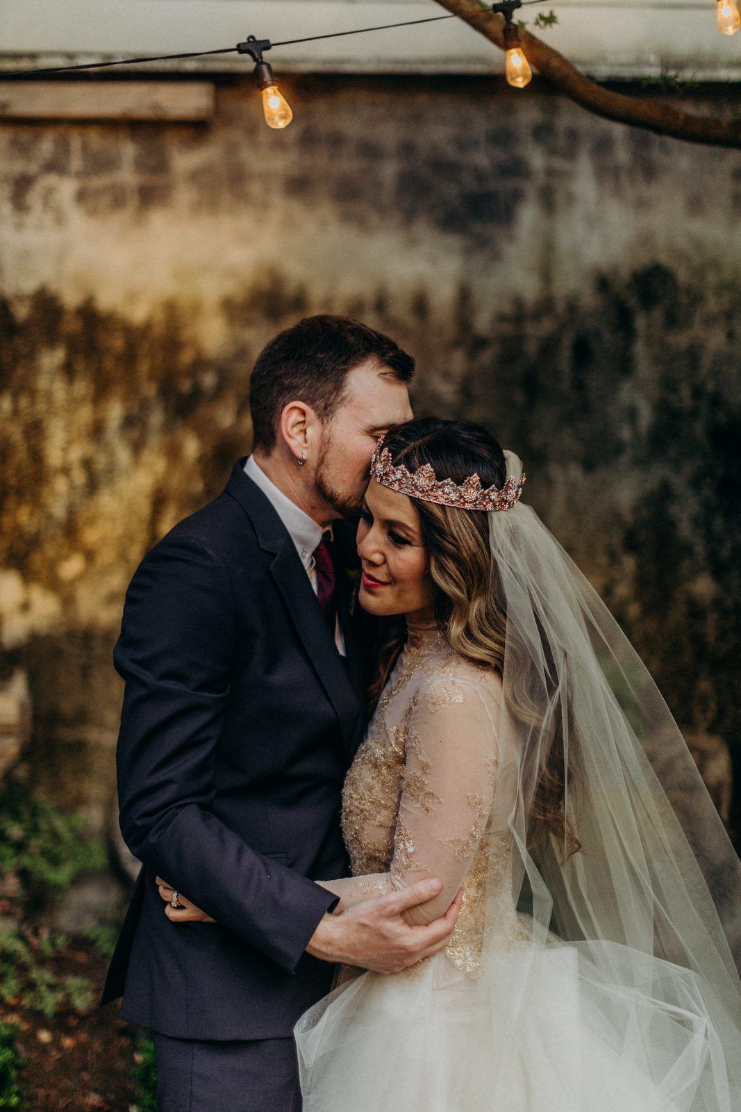The couple in a courtyard under a string of lights, the groom resting his head against the bride's as she leans into him in her crown and veil.