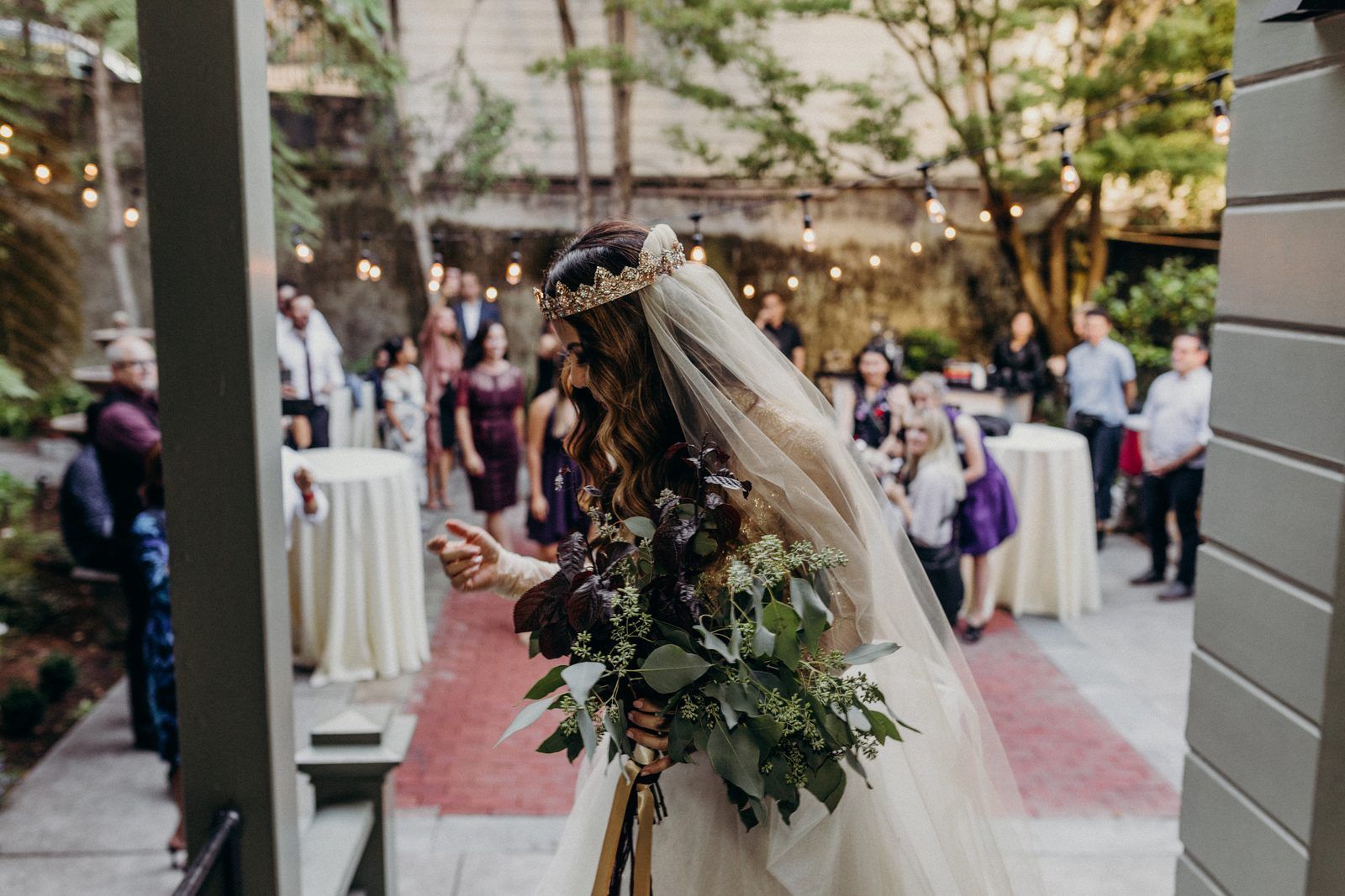 The bride in her crown and veil holding a green bouquet in the courtyard garden, guests standing among string lights and tables beyond.