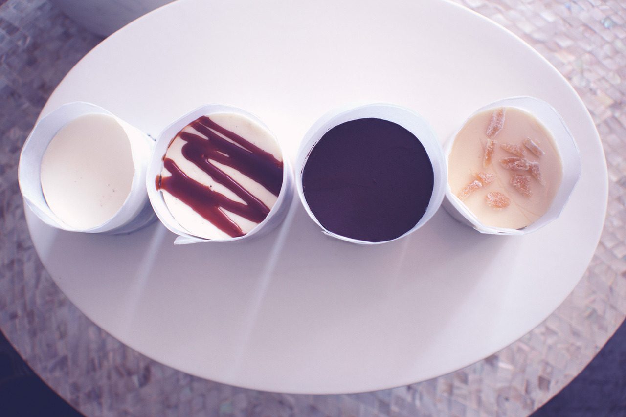 Overhead view of four cake tasting cups on a white oval plate: vanilla cream, caramel drizzle, dark chocolate and candied ginger.