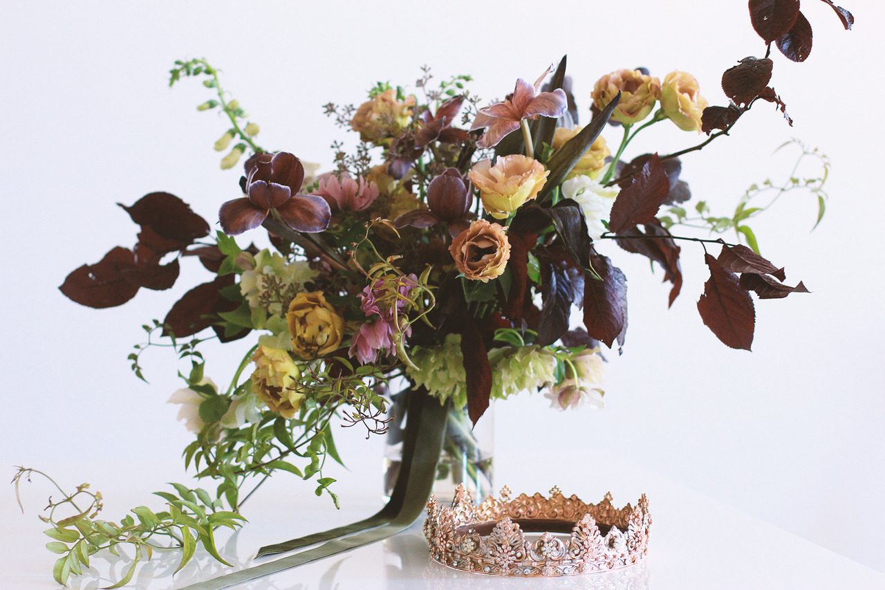 A rose gold bridal crown beside a dark, moody bouquet of burgundy foliage, tulips and lisianthus in a vase.