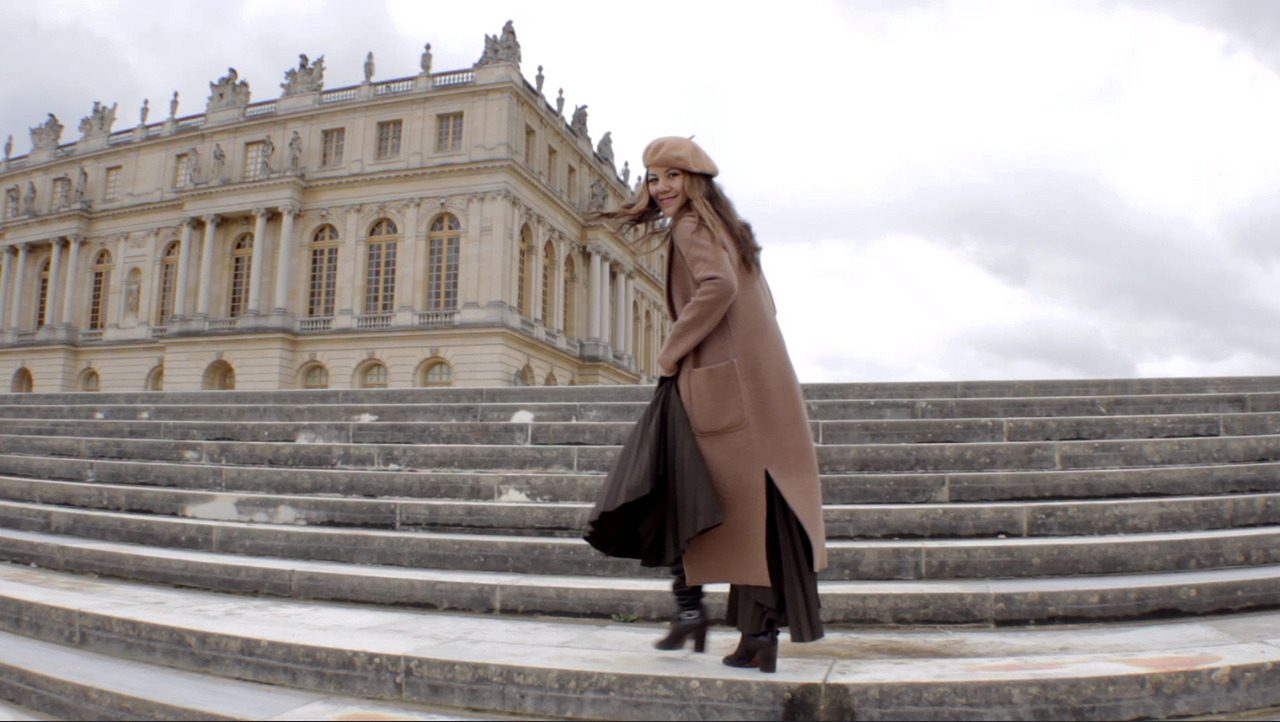 A woman in a camel coat and beret on the steps of the Palace of Versailles.